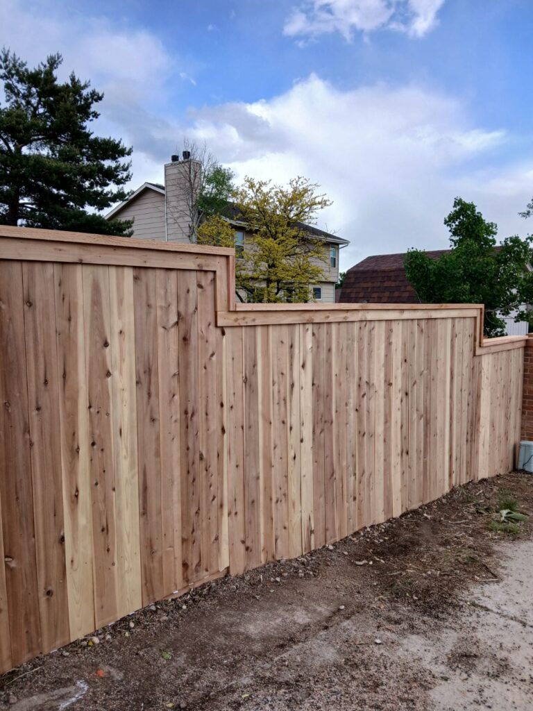 A newly constructed wooden privacy fence with a stepped top design runs alongside a dirt path, with houses and trees in the background.