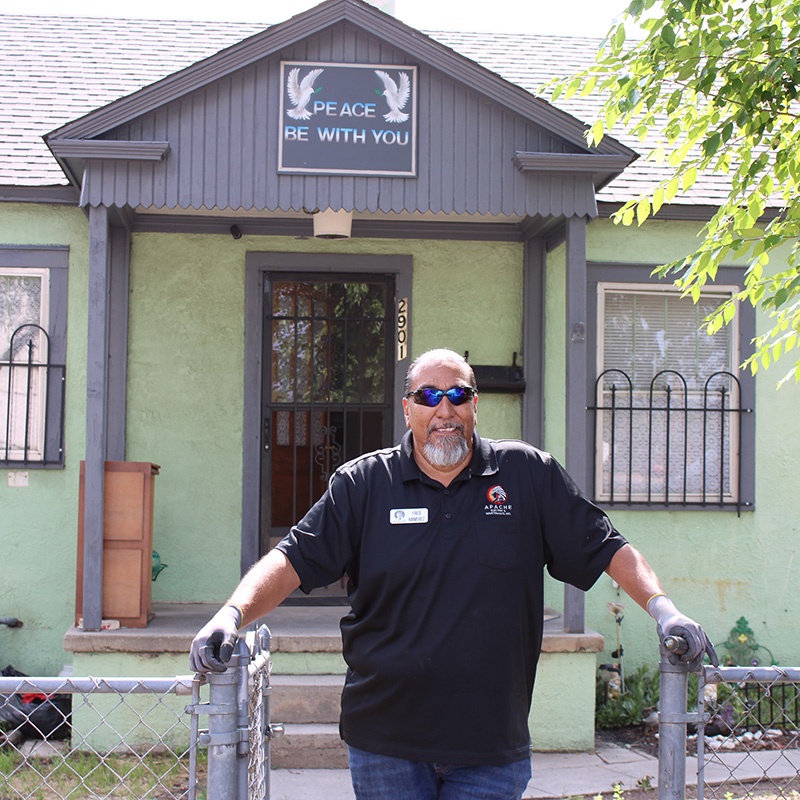 A man wearing sunglasses and gloves stands at a gate in front of a green house with a sign above the door that reads "PEACE BE WITH YOU," welcoming guests from his business networking group.