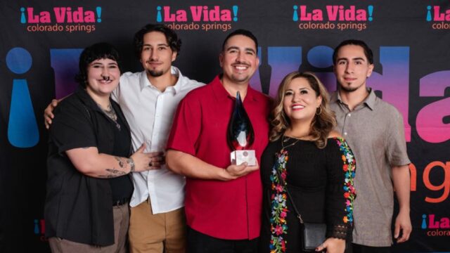 Five people stand together smiling in front of a "¡La Vida! Colorado Springs" backdrop; the person in the center holds a glass award.