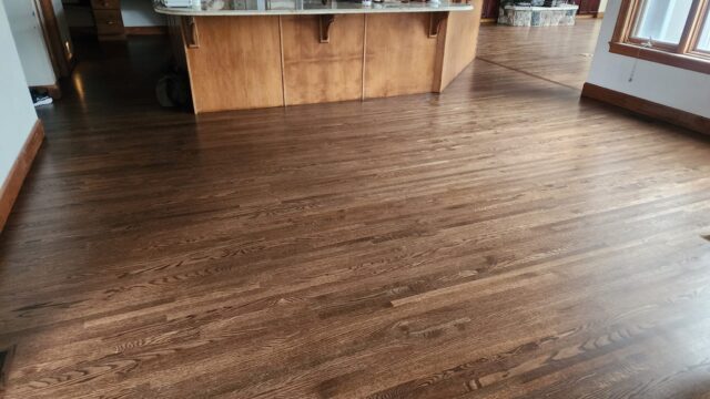 A large room with dark-stained hardwood floors, a wooden kitchen island with a marble countertop, and natural light from a window.