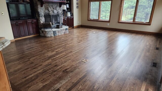Living room with polished wooden floors, a stone fireplace, built-in cabinets, large windows, and trees visible outside.