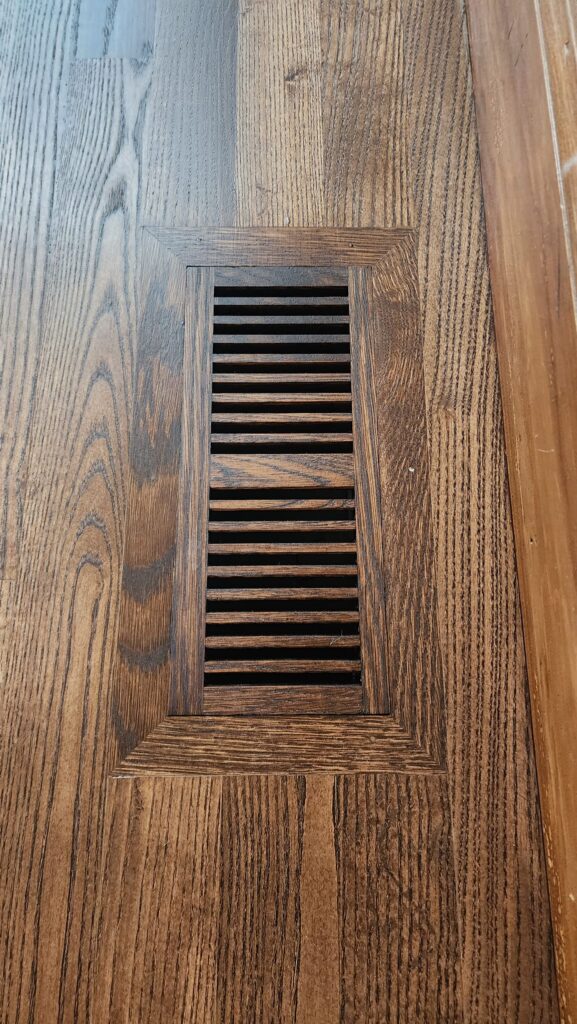 A wooden floor vent cover is seamlessly integrated into hardwood flooring near a wooden baseboard.