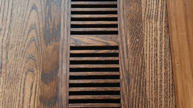 A wooden floor vent cover is seamlessly integrated into hardwood flooring near a wooden baseboard.