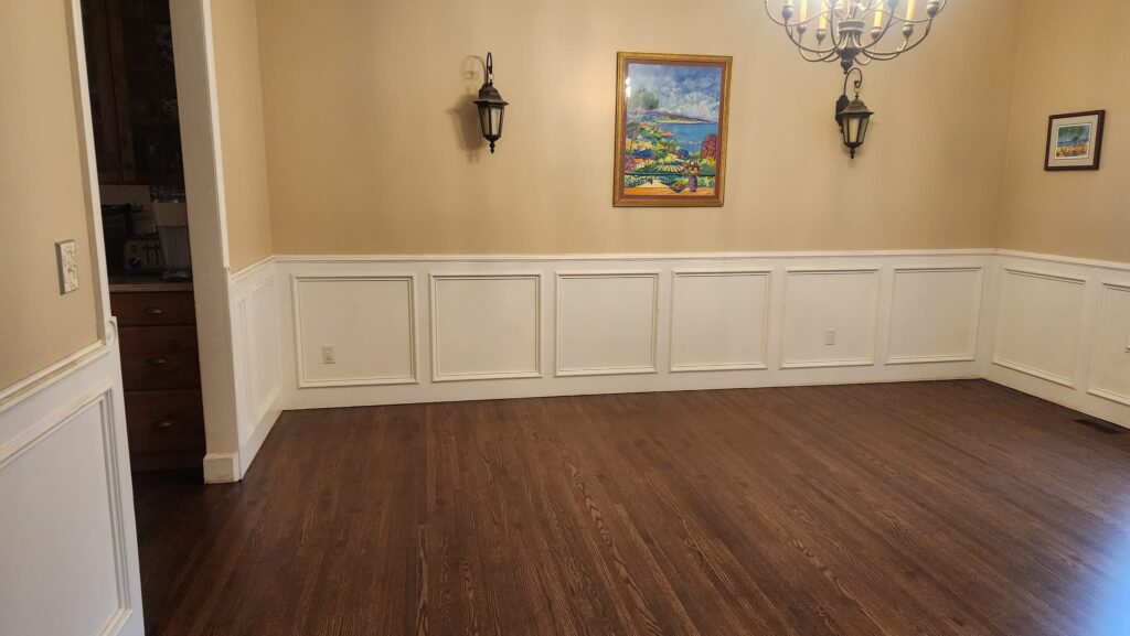 Empty dining room with hardwood floors, beige walls, white wainscoting, wall sconces, a chandelier, and colorful framed artwork on the wall.