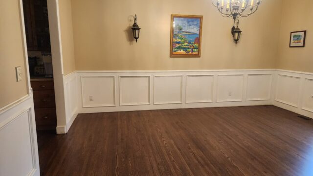 Empty dining room with hardwood floors, beige walls, white wainscoting, wall sconces, a chandelier, and colorful framed artwork on the wall.