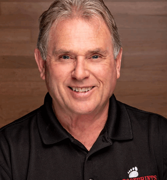A middle-aged man with gray hair, wearing a black polo shirt with the "Footprints Floors" logo, smiles in front of a wooden background.