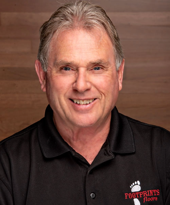 A middle-aged man with gray hair, wearing a black polo shirt with the "Footprints Floors" logo, smiles in front of a wooden background.