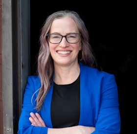 A woman with long, grayish hair and glasses, wearing a blue blazer and black top, stands with arms crossed, smiling at the camera.
