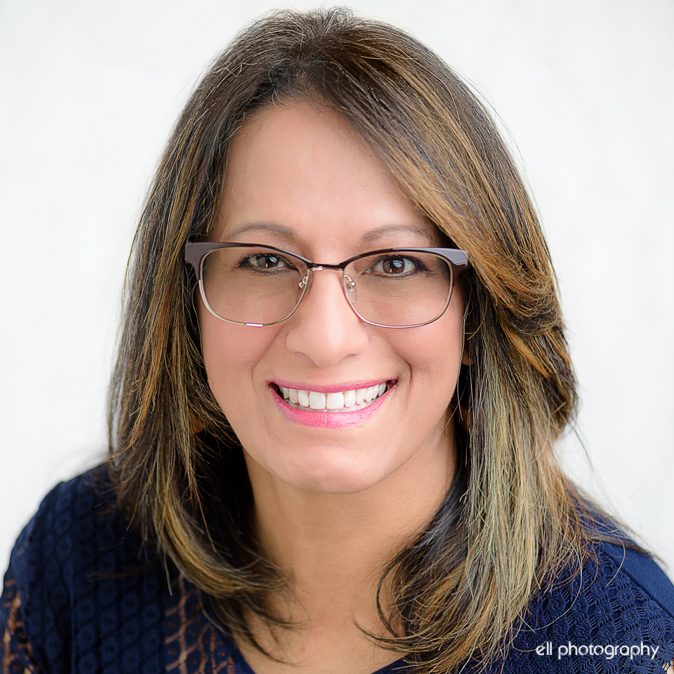 A woman with straight brown hair and glasses smiles at the camera, wearing a dark blue patterned top against a white background.