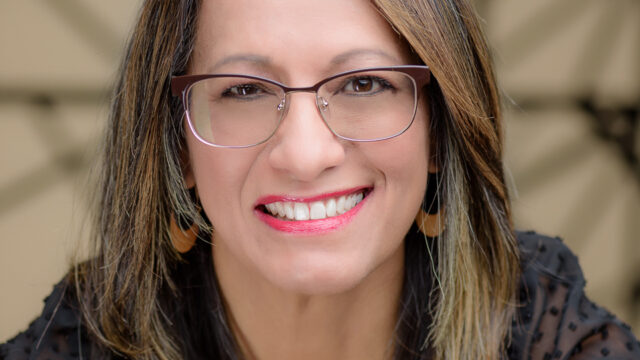 Woman with straight, shoulder-length hair and glasses, wearing a black top, smiling at the camera in front of a neutral patterned background.